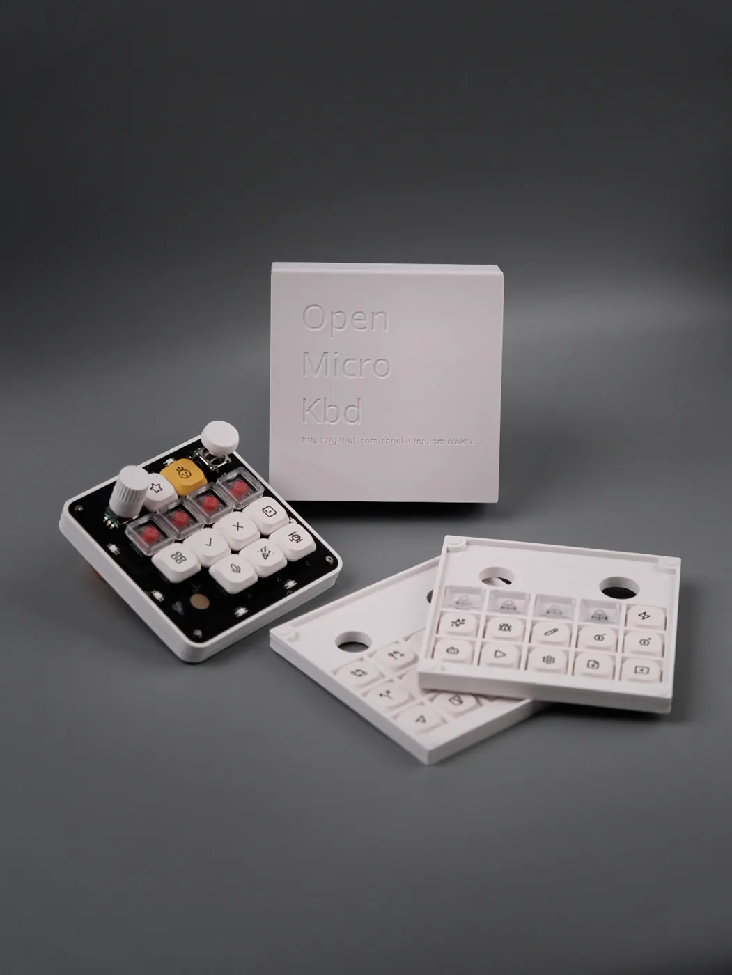 The assembled board in its white printed case, beside the embossed OpenMicroKbd packaging box and two printed trays of alternate keycaps