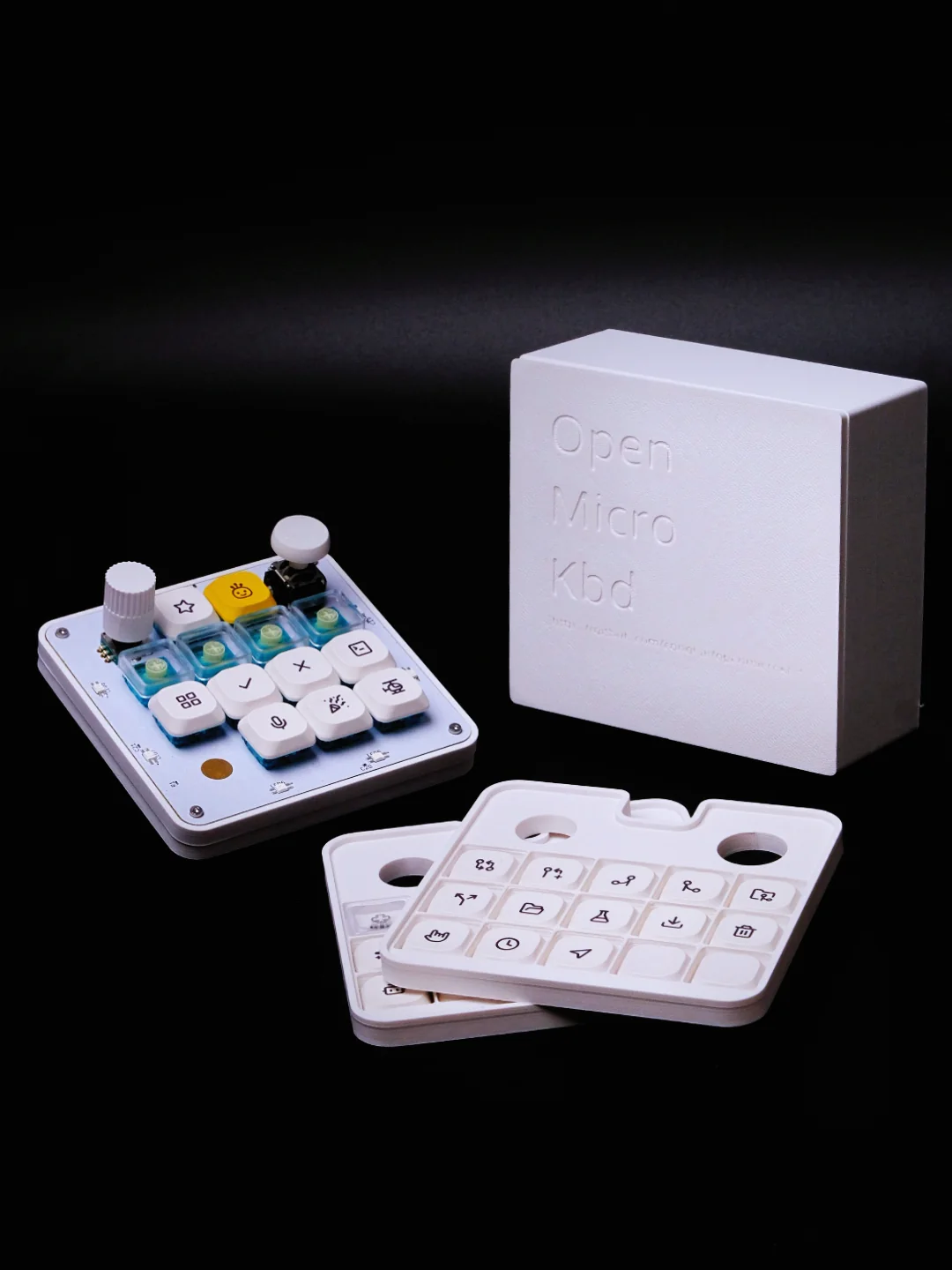 The white OpenMicroKbd macropad with its rotary encoder, joystick, keycaps, and 3D-printed packaging on a black background