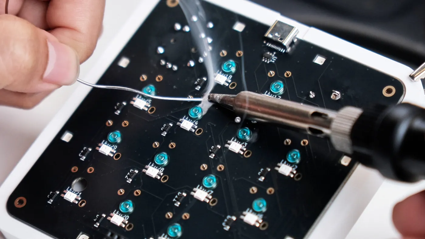 Macro view of the board's underside during hand assembly: a soldering iron at one joint, surrounded by teal reverse-mount LED packages silkscreened LED17 through LED29